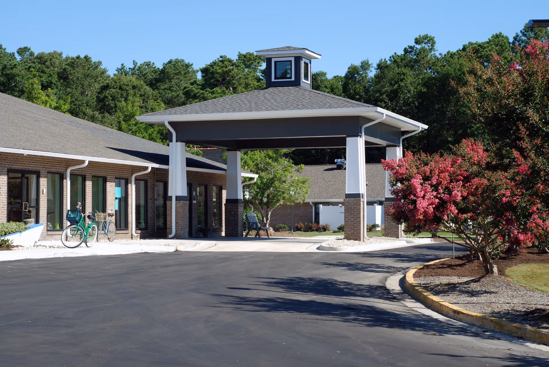 Entrance of a single-story brick senior living facility with a covered porte-cochere, bicycles near the door, and a flowering tree by the driveway.