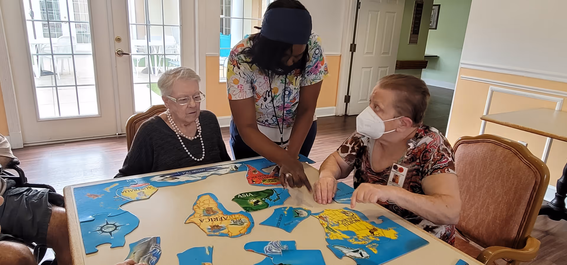 Two elderly women and a caregiver wearing a mask are sitting around a table working on a large puzzle of a world map in a well-lit room with glass doors leading outside.