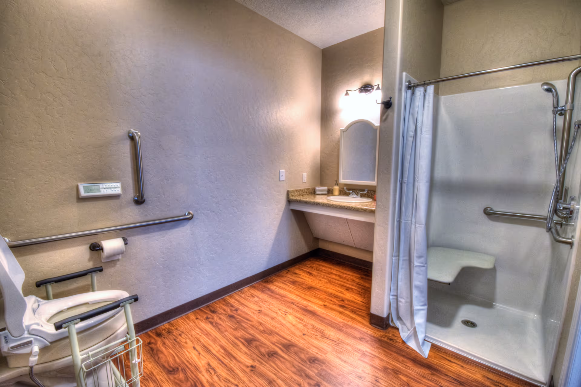 Accessible bathroom with wooden flooring, a shower area with a built-in seat and grab bars, a white shower curtain, a wall-mounted sink with a mirror and light above it, and a raised toilet with armrests and grab bars on the wall.