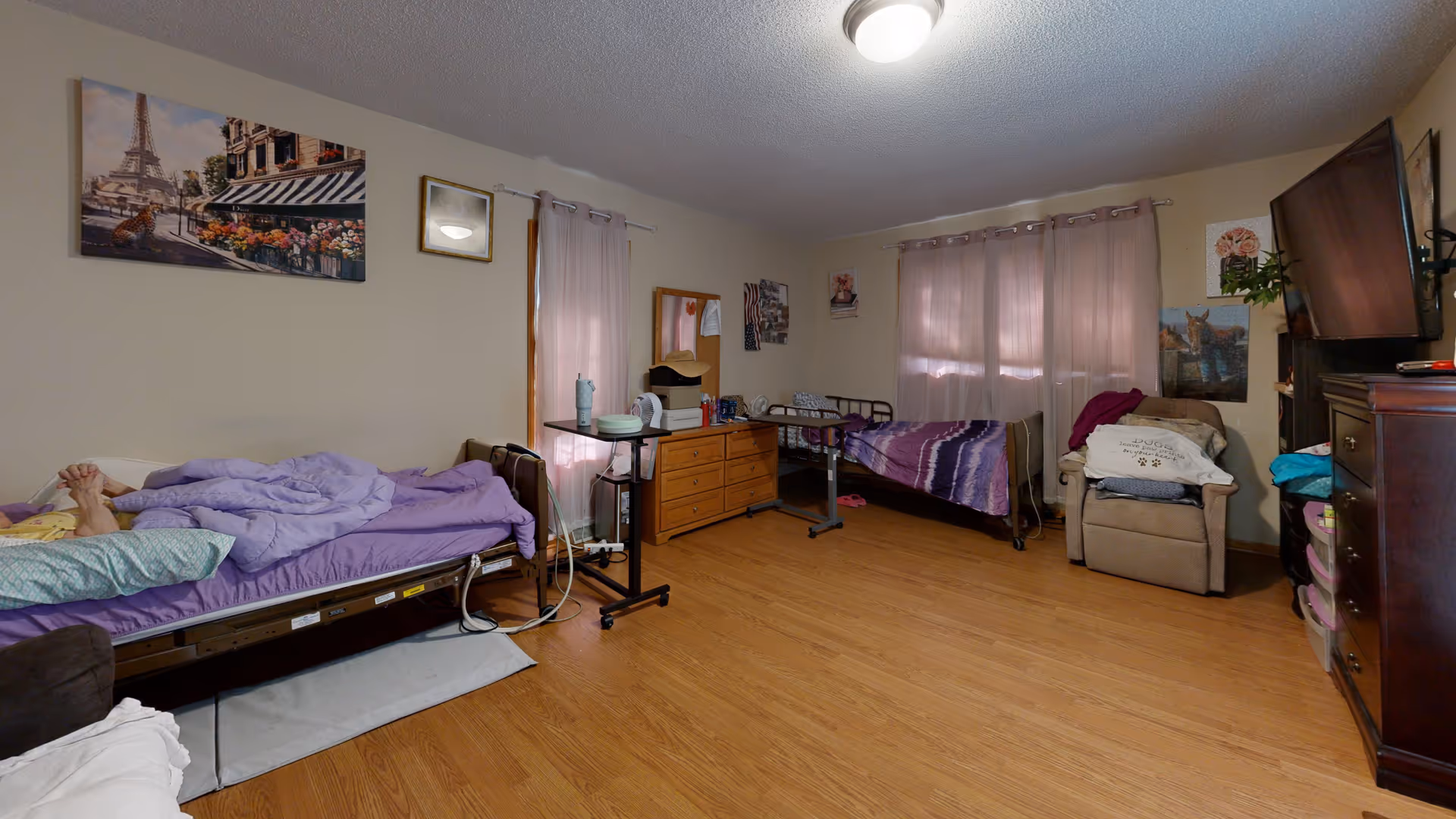 A senior living bedroom with two hospital-style beds, each with purple bedding. The room has wooden flooring, a dresser with a mirror, a reclining chair with folded blankets, a wall-mounted TV, and several framed pictures on the walls. Light pink curtains cover the windows.