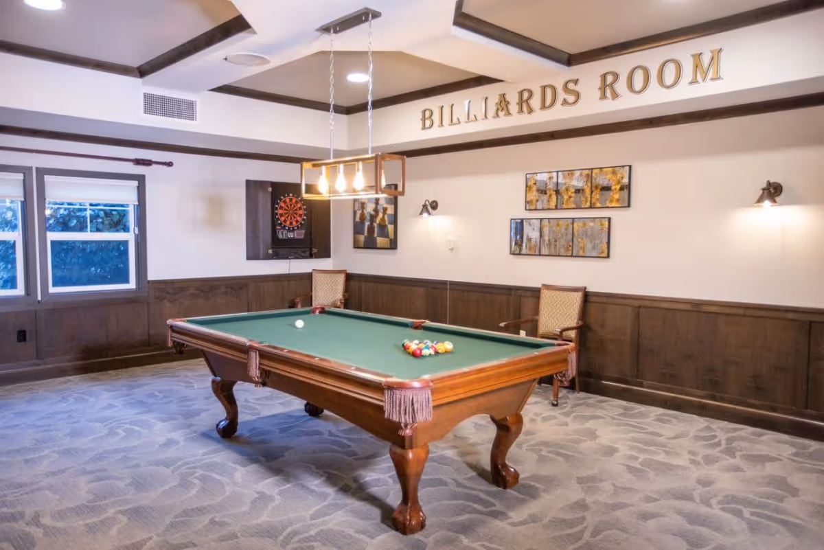 A billiards room with a green felt pool table in the center, a dartboard mounted on the wall, two wooden chairs with cushions, decorative wall art, and a ceiling light fixture above the pool table. The room has carpeted flooring and wood paneling on the lower half of the walls. The words 'BILLIARDS ROOM' are displayed prominently on the wall.