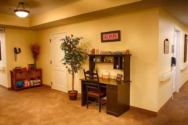 A cozy hallway area in an assisted living facility with beige walls and carpet. There is a dark wooden desk with a chair, decorated with books, a basket, and framed photos. Above the desk is a framed sign that says 'HOME'. To the left, there is a small wooden shelf with baskets and books, a wall-mounted phone, and a tall plant in a pot. The hallway extends to the right with doors and handrails along the walls.