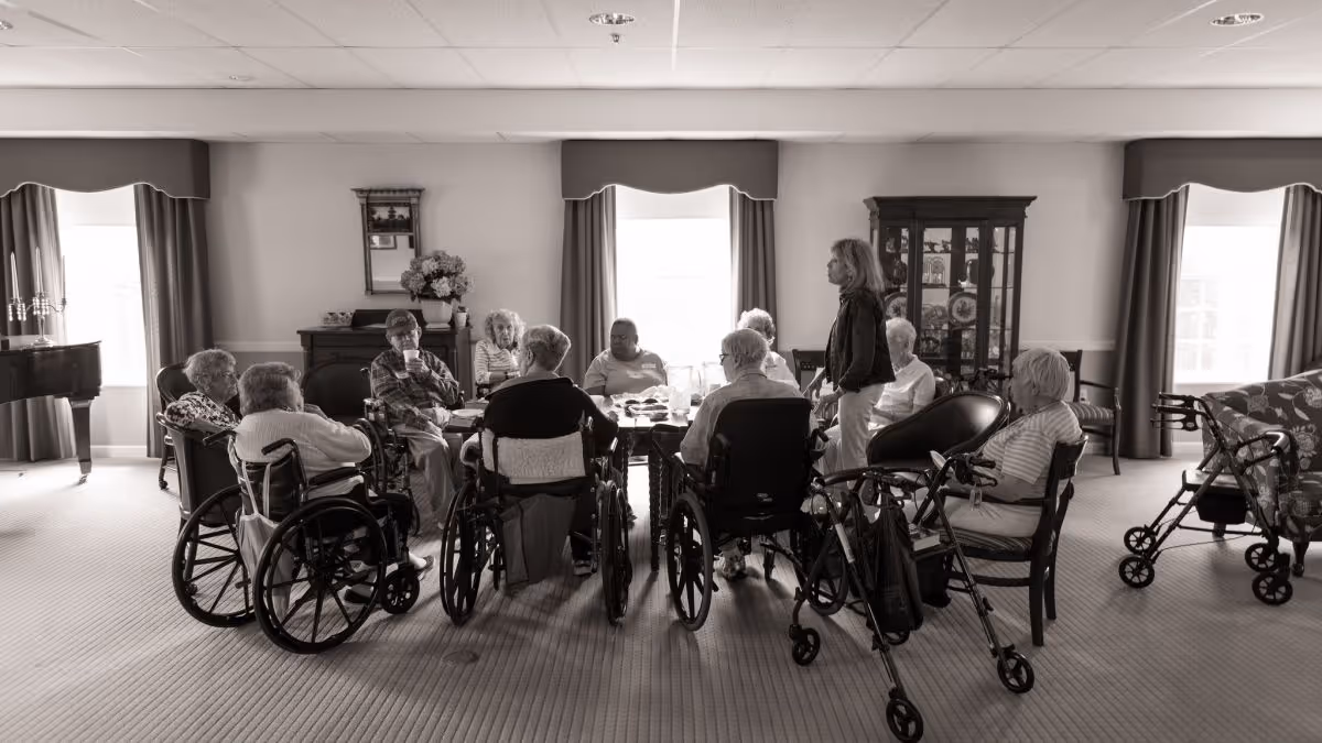 A group of elderly residents, some in wheelchairs, gathered around a table in a bright common room with a staff member standing nearby.