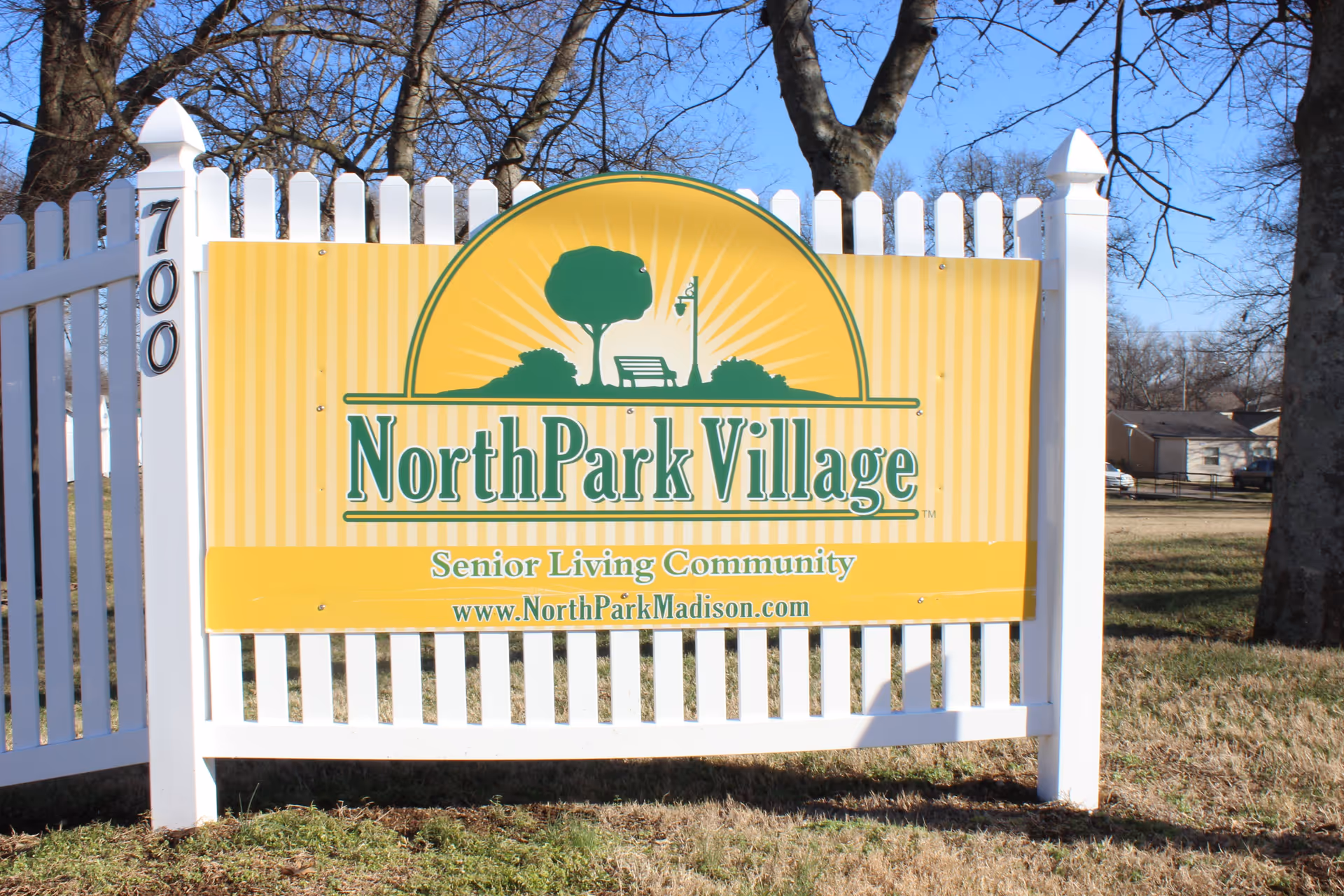A yellow and green sign for North Park Village Senior Living Community mounted on a white picket fence with the address number 700 on the left post. The sign features a graphic of a tree, a bench, and a lamp post with a sunburst background. The website www.NorthParkMadison.com is displayed at the bottom of the sign. Trees and houses are visible in the background under a clear blue sky.