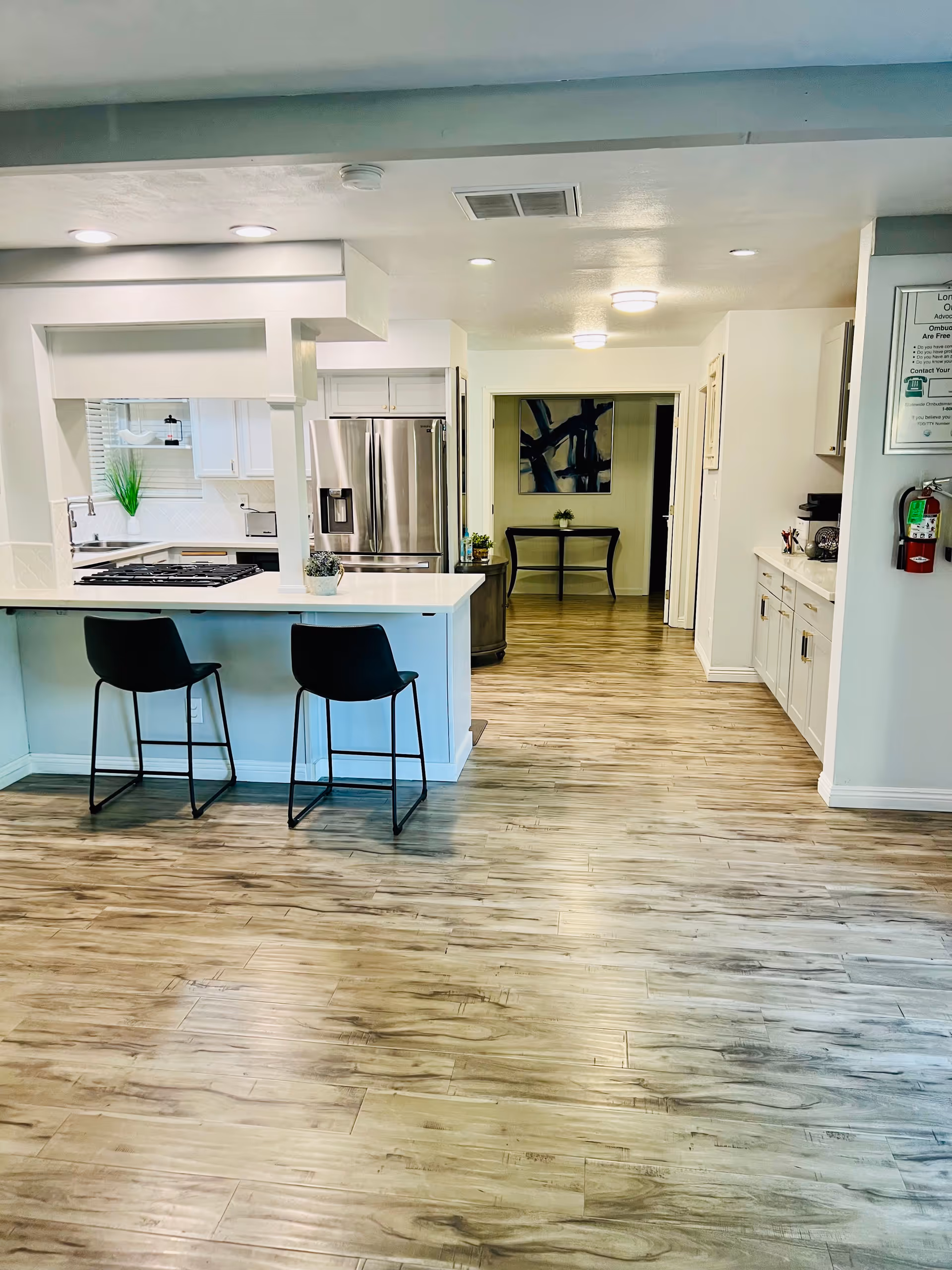Interior view of a modern kitchen area with light wood flooring, white cabinetry, a stainless steel refrigerator, and a kitchen island with two black bar stools. The space is well-lit with recessed ceiling lights and a small table with a plant is visible in the background hallway.