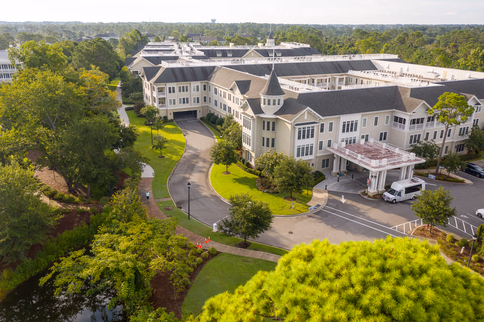 Aerial view of Bradley Creek Health Center at Carolina Bay at Autumn Hall, showing a large multi-story building with a turret, surrounded by green trees and landscaped grounds. A driveway leads to the main entrance where a white van is parked. The area is lush with greenery and a small pond is visible in the lower left corner.