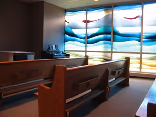 Interior of a small chapel or meditation room with wooden pews facing a large stained glass window featuring abstract blue, white, and red wave patterns.