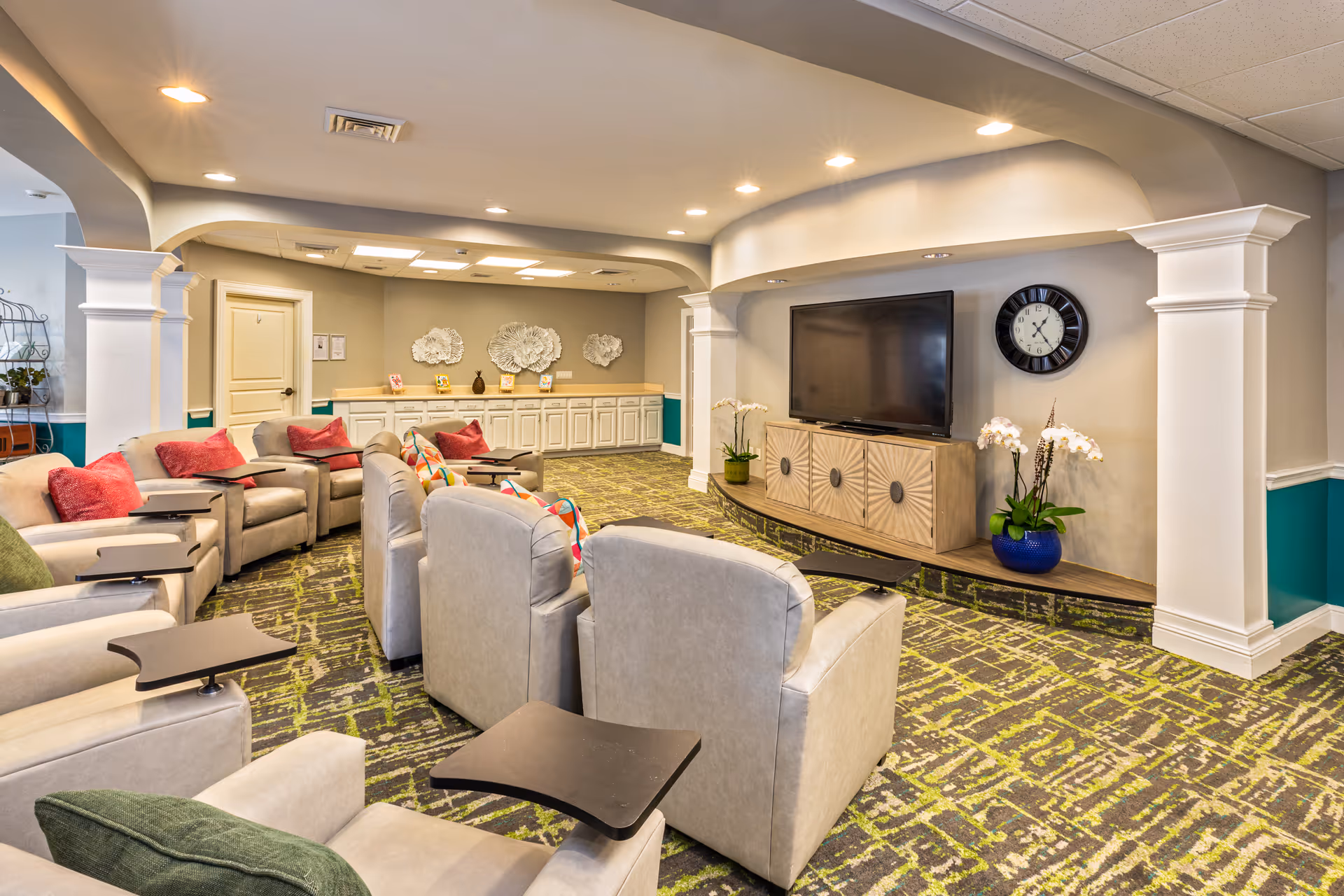 A cozy living room area in an assisted living facility with multiple beige armchairs arranged in rows facing a large flat-screen TV mounted on a wooden cabinet. The room features a patterned green and gray carpet, recessed ceiling lights, decorative wall art, and a wall clock. There are colorful throw pillows on the chairs and potted plants near the TV cabinet.