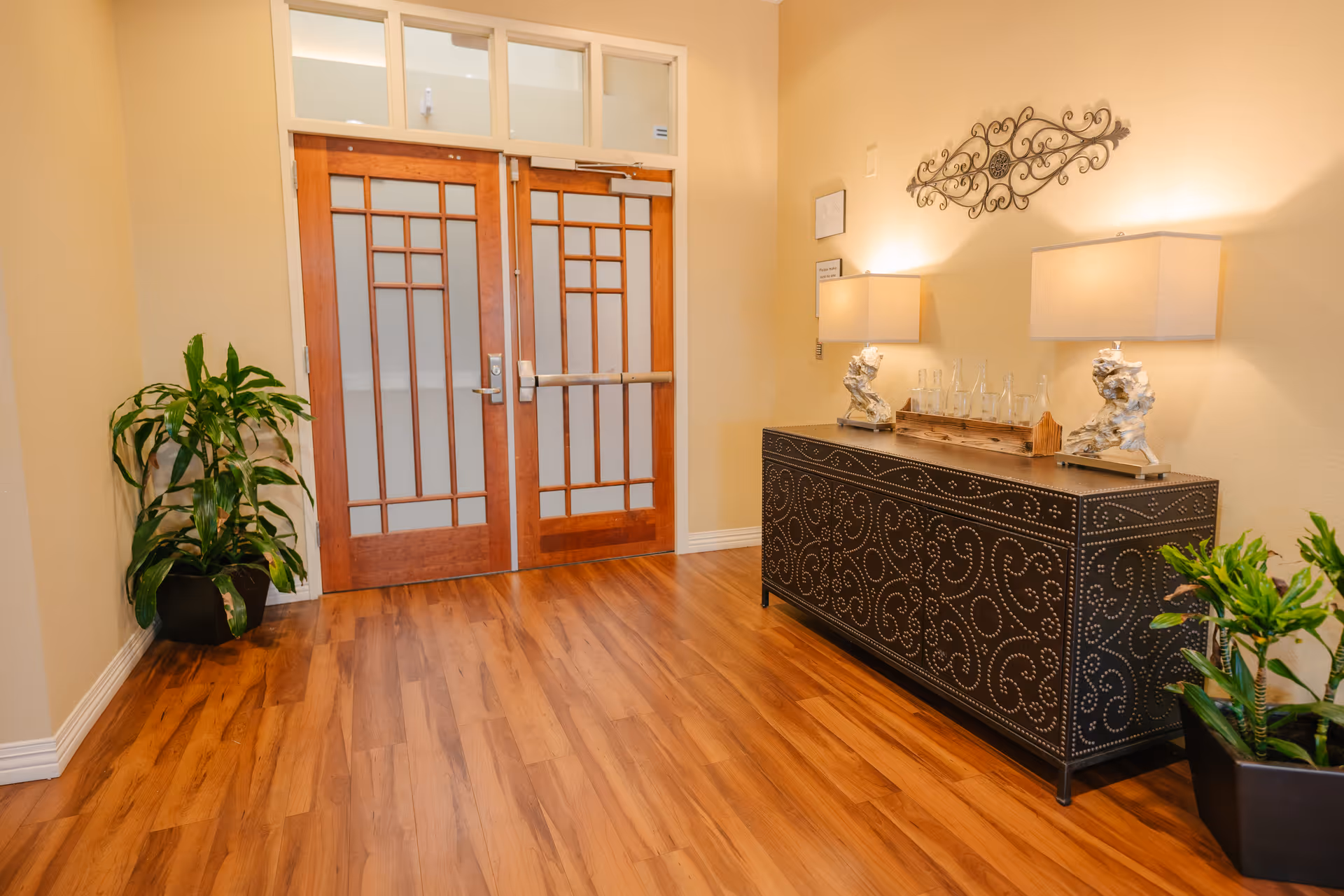 Interior hallway with wooden double doors featuring glass panels, a decorative black sideboard with intricate patterns, two table lamps, a tray with glass bottles, wall art above the sideboard, and potted plants on either side of the hallway. The floor is wooden and the walls are painted light beige.