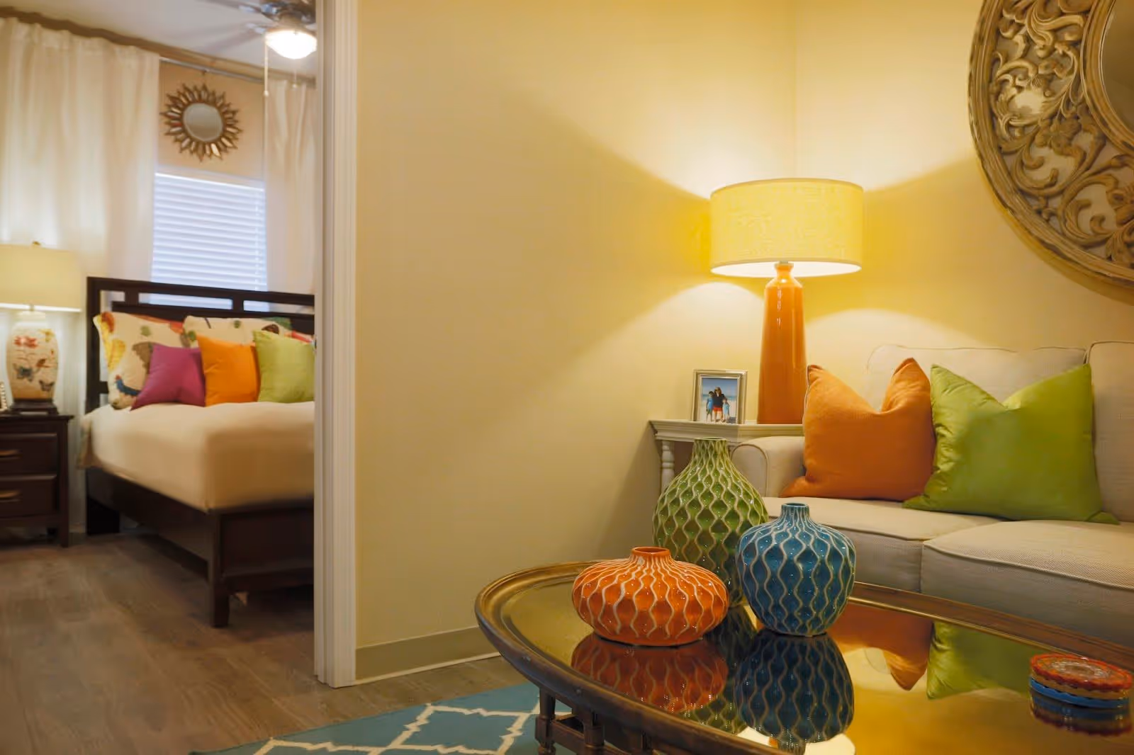 A cozy living room area with a beige sofa adorned with orange and green throw pillows, a side table with a lamp and a framed photo, and a glass coffee table with three decorative vases in orange, green, and blue. In the background, a bedroom is visible with a bed featuring colorful pillows and a bedside table with a lamp, under a window with white curtains.