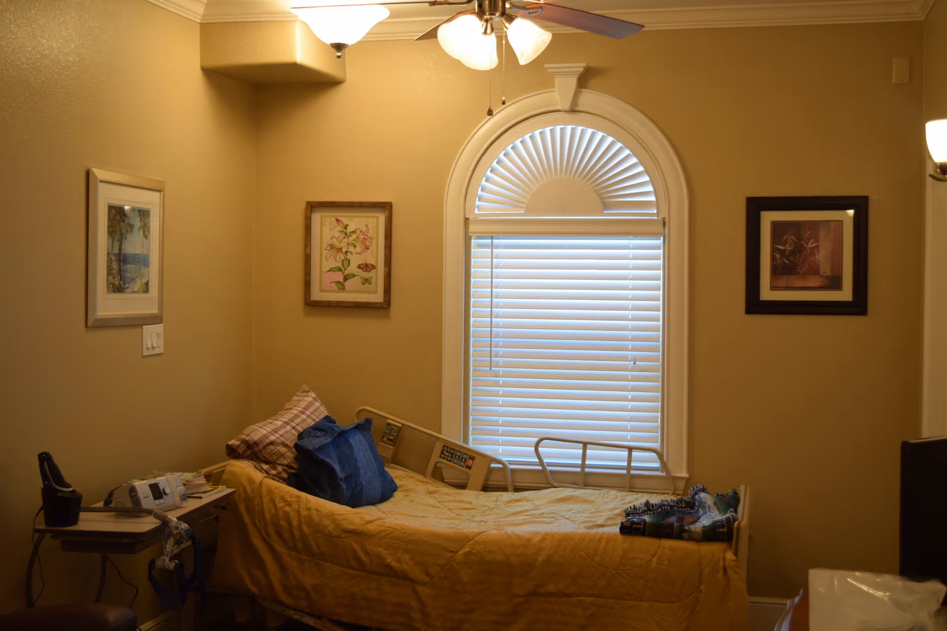 A small bedroom in an assisted living facility with a single bed covered in a yellow blanket and pillows. The bed has side rails and is positioned against a wall with a window featuring white blinds and an arched top. There are three framed pictures on the beige walls, a ceiling fan with lights, and a small table with medical equipment and personal items next to the bed.