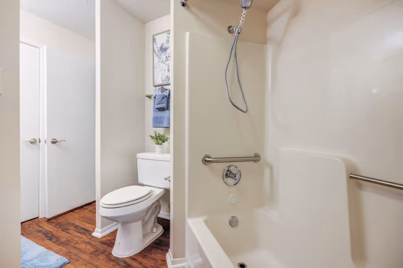 A bathroom featuring a white toilet with a small potted plant on the tank, a towel rack with blue towels, and a shower-tub combo with built-in seating and grab bars. The floor has wood-like flooring and there are two white doors visible in the background.