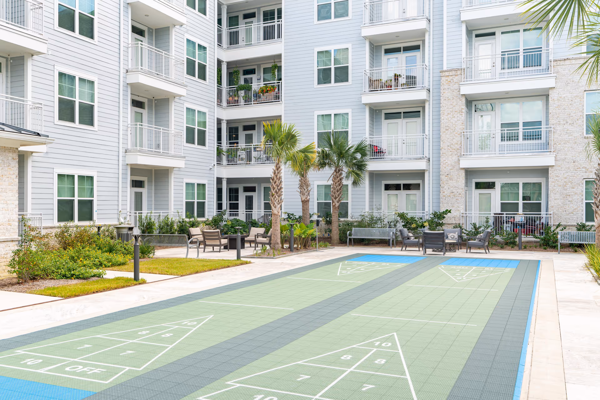 Outdoor courtyard area of a senior living facility featuring two shuffleboard courts, surrounded by palm trees, benches, chairs, and tables. The background shows a multi-story residential building with balconies and large windows.