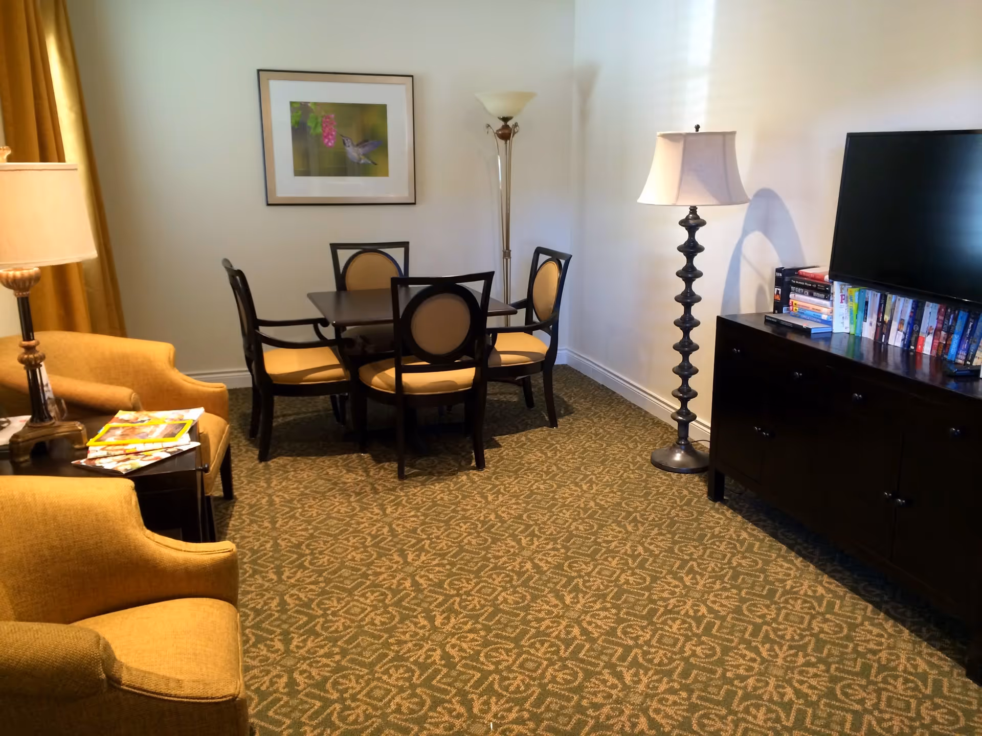A cozy living area with a small dining table surrounded by four chairs with beige cushions. To the left, there are two mustard-yellow armchairs next to a side table with magazines and a lamp. On the right side, there is a dark wooden TV stand with a flat-screen television and a collection of DVDs or books. The room has patterned carpet flooring, a floor lamp, and a framed picture of a hummingbird on the wall.