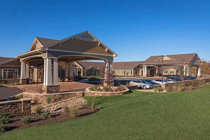 Front exterior of a single-story care facility with a covered porte-cochère, landscaped lawn, and parked cars under a clear blue sky.