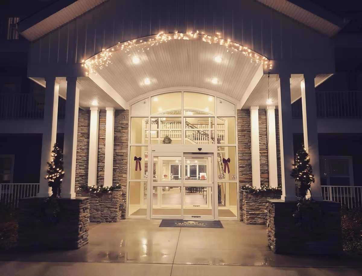 Night view of the entrance to The Chelsea at Brookhaven facility, featuring a covered porch with stone pillars decorated with small Christmas trees and string lights. The glass doors reveal a well-lit interior with visible stairs and seating area.