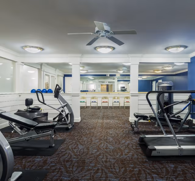 Well-lit indoor gym with treadmills, weight benches, exercise machines, a ceiling fan, and mirrored walls.