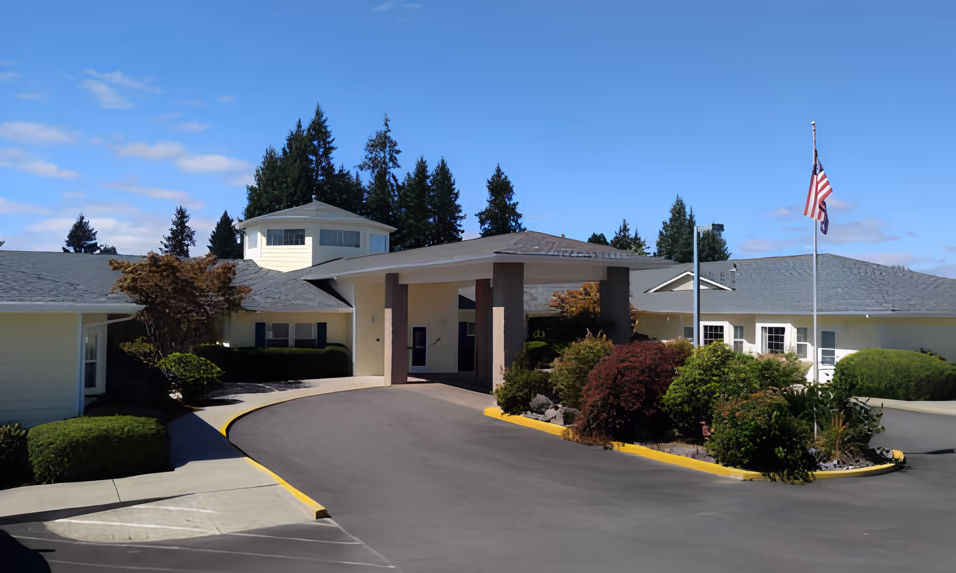 Front entrance of a single-story assisted living facility with a covered drive, landscaped island, and an American flag flying on a pole.