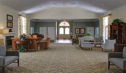 Spacious, carpeted communal lounge with sofas and armchairs arranged around small tables and double doors at the far end.