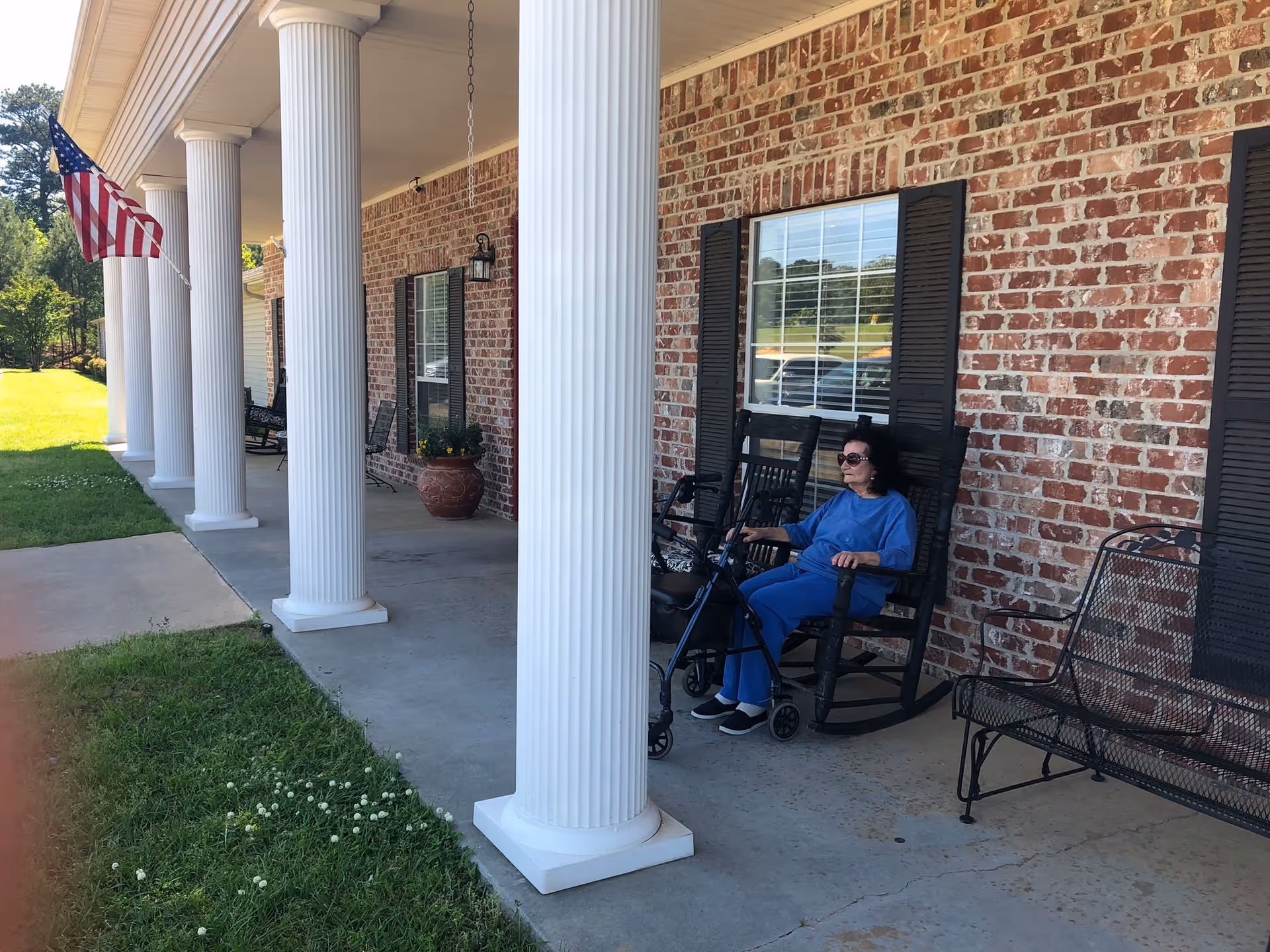 A woman wearing blue clothing and sunglasses sits on a black rocking chair on a covered porch with white columns and a brick wall. There is a black metal bench nearby and an American flag hanging from the porch. The porch overlooks a grassy area.