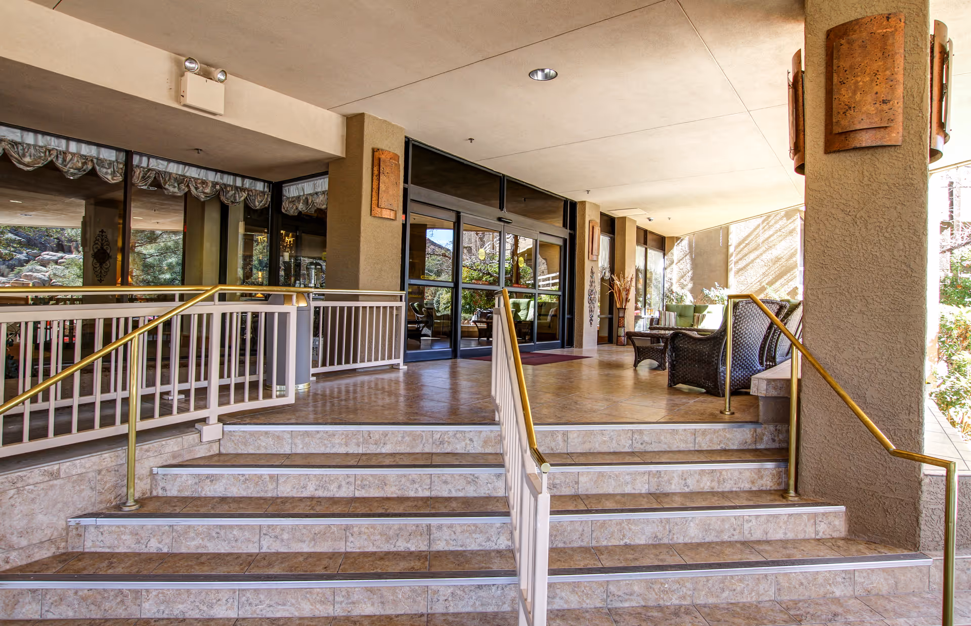 Entrance area of a senior living facility with tiled steps leading up to glass double doors. There are handrails on both sides of the steps and a ramp on the left side. The covered porch area has seating with wicker chairs and cushions, and large windows reflecting the outdoor scenery.