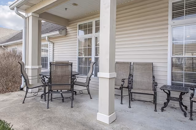 Outdoor patio area with a round metal table and four chairs, two additional chairs and a small side table against the beige siding of a building with windows.