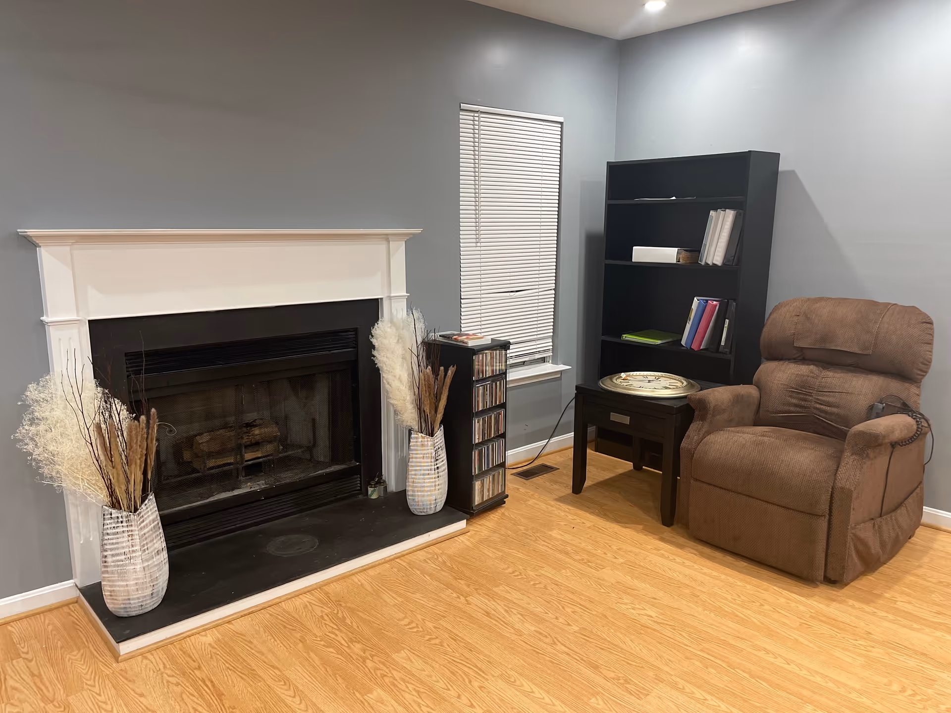 A cozy living room corner featuring a brown recliner chair with a remote control, a small dark wooden side table with a large decorative clock on top, a black bookshelf with binders and books, a window with closed blinds, and a fireplace with a white mantel flanked by two vases filled with dried decorative plants. The room has light wood flooring and gray walls.