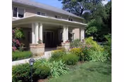 Exterior view of a building with a covered porch supported by columns, surrounded by green grass, bushes, and flowering plants under a clear sky.
