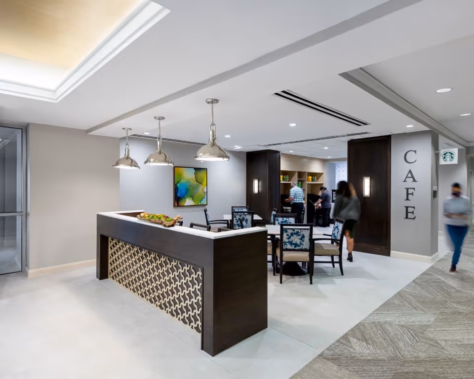 Interior view of a modern cafe area in a senior living facility with a dark wood counter featuring a decorative front panel, three hanging pendant lights, several tables and chairs with patterned upholstery, and people walking and standing in the background. The word 'CAFE' is displayed vertically on a wall near the entrance.
