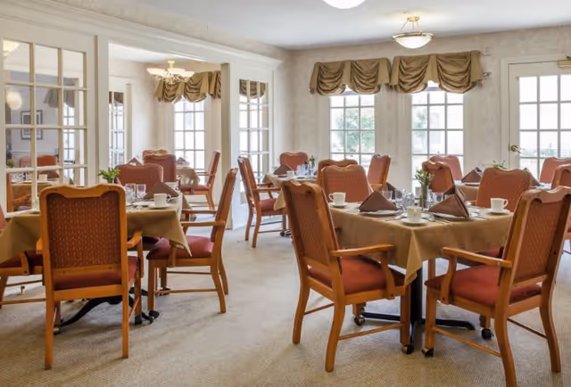 A bright dining room with multiple tables covered in beige tablecloths, each set with cups, glasses, napkins, and silverware. The chairs have wooden frames with red cushioned seats and backs. Large windows with brown valances allow natural light to fill the room.