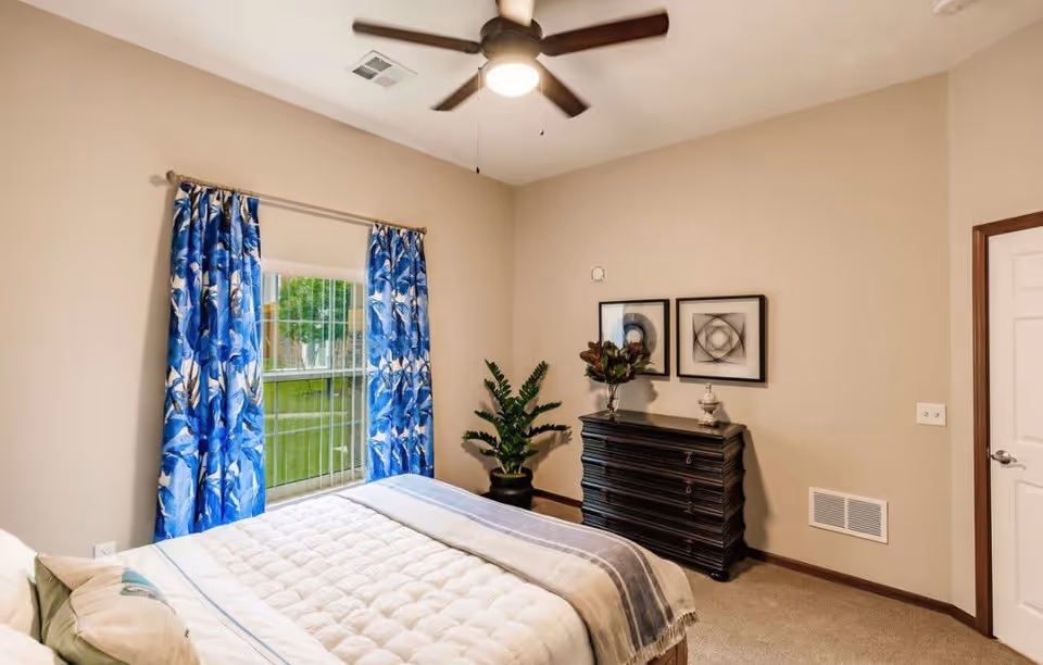 A neatly staged bedroom with a bed, blue floral curtains at a window, a ceiling fan, dark dresser and potted plant.