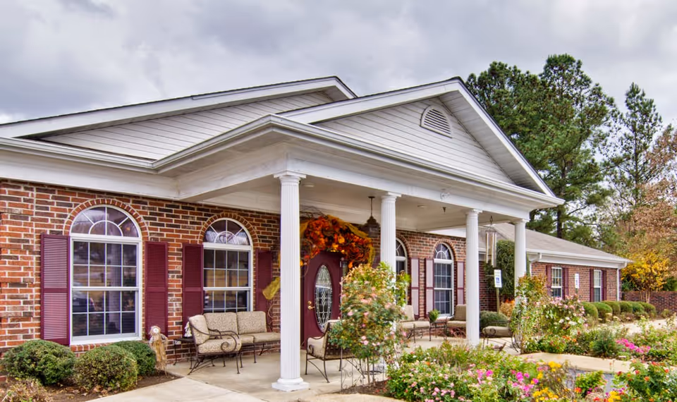 Exterior view of a single-story brick building with white columns supporting a covered entrance. The building has arched windows with maroon shutters and a maroon door decorated with a fall-themed wreath. There are benches on the porch and landscaped bushes and flowers in front of the building under a cloudy sky.