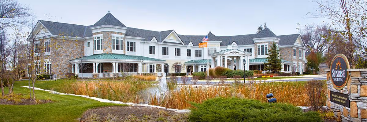 Exterior view of a large, two-story senior living facility with stone and white siding, multiple windows, and a covered entrance. The building is surrounded by landscaped grounds with grass, shrubs, a small pond, and trees. A sign near the entrance reads 'Sunrise Senior Living.'