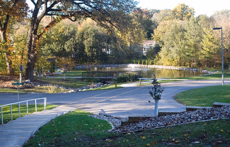 A peaceful outdoor scene at Meth-Wick Community featuring a paved road curving around a small pond with a fountain in the center. There are trees with autumn foliage surrounding the area, a small bridge over the pond, a street lamp, and a set of stairs with a handrail leading down to the road.