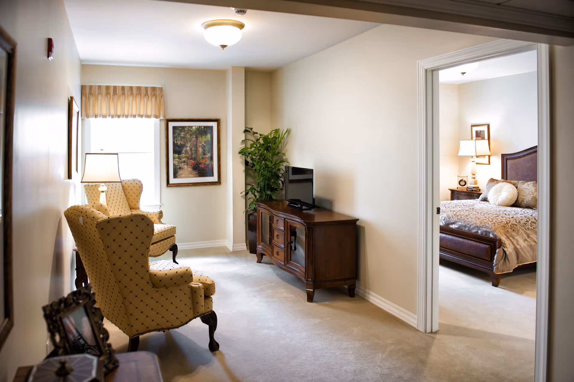 A cozy sitting area with two yellow patterned armchairs, a wooden TV stand with a flat-screen TV, a potted plant, and a framed painting on the wall. Through an open doorway, a bedroom with a bed, nightstand, lamp, and framed pictures is visible.