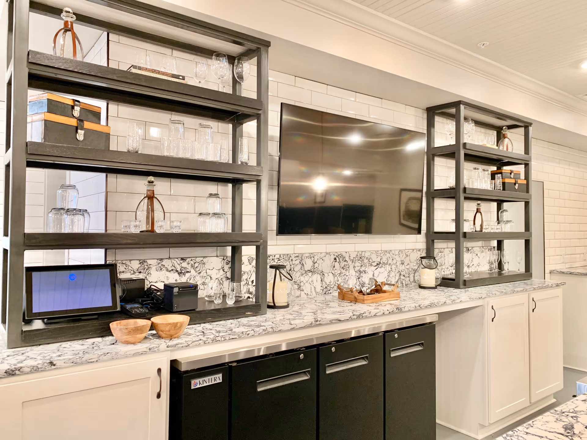 Marble countertop and open shelving stocked with glassware and a wall-mounted TV in a bright kitchen/bar area.