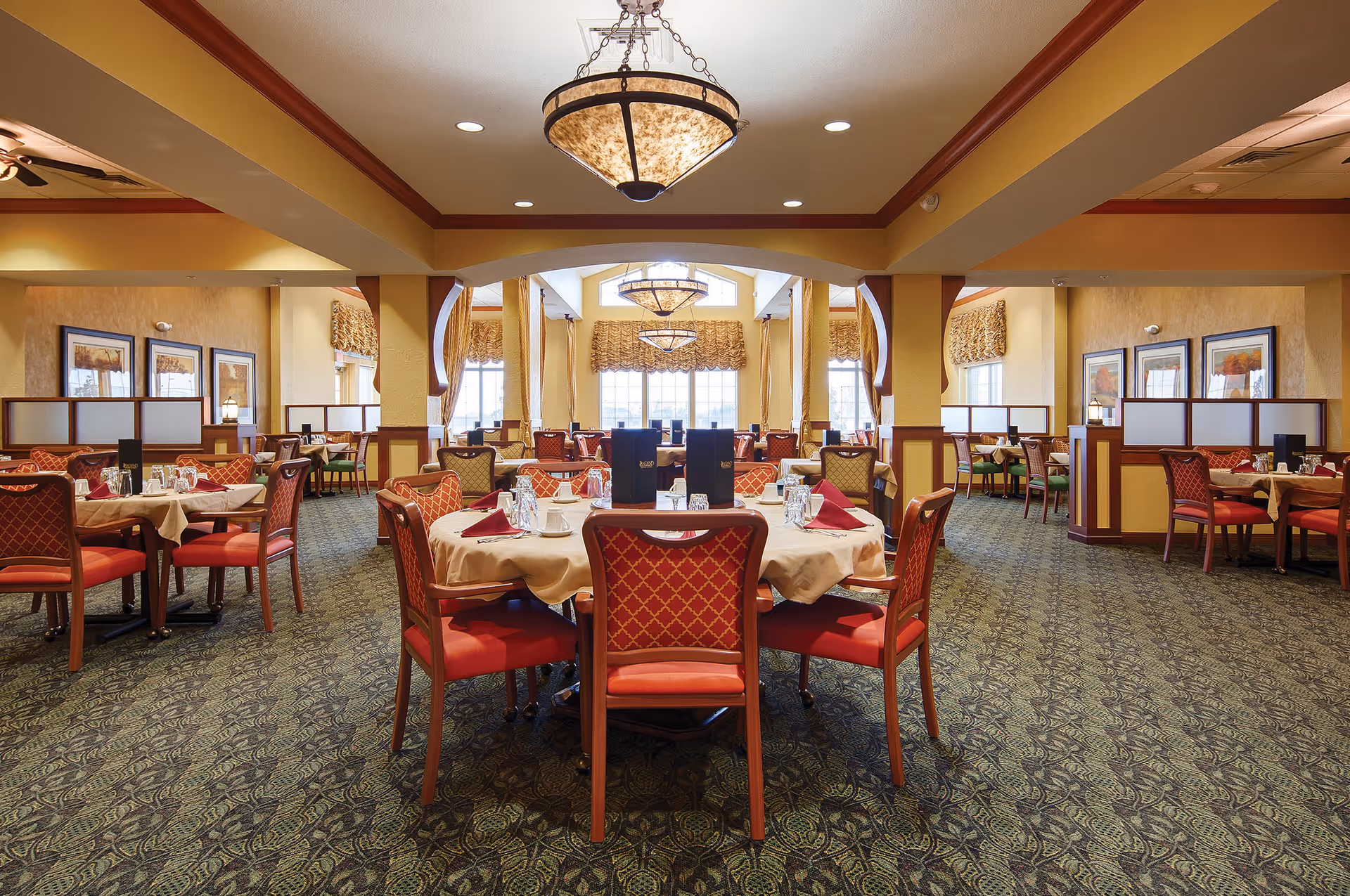 A spacious and elegant dining room with round tables covered in beige tablecloths, set with glasses, cups, and red napkins. The room features red cushioned chairs with patterned backs, large windows with decorative curtains, framed artwork on the walls, and warm lighting from ceiling fixtures.
