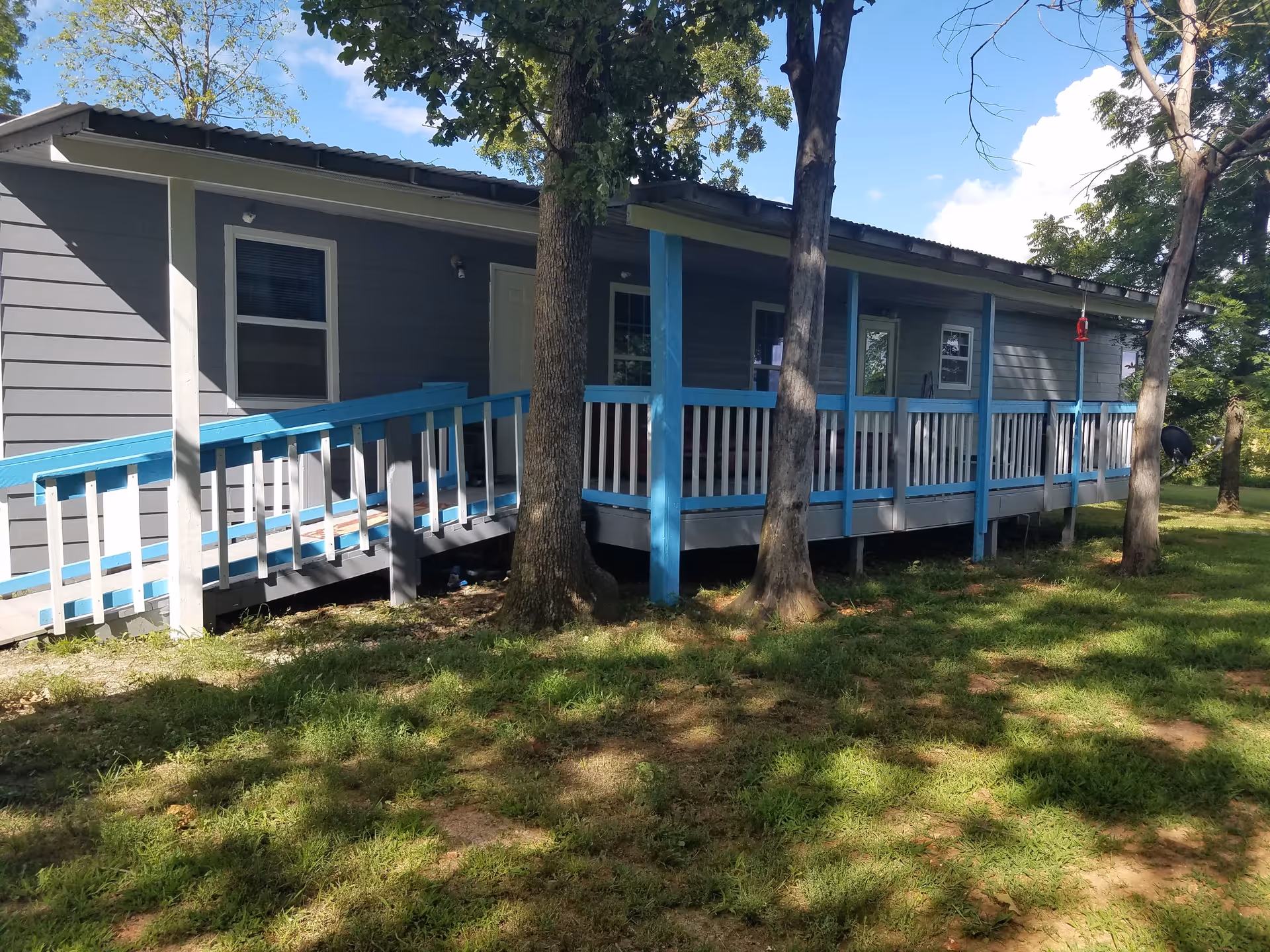 Exterior view of a single-story gray building with a blue and white ramp and porch railing surrounded by trees and grass under a partly cloudy sky.