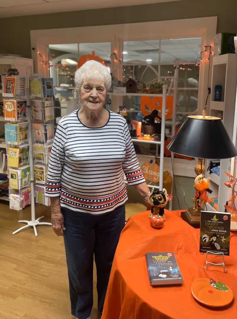 An elderly woman with white hair wearing a striped shirt and dark pants stands indoors next to a table covered with an orange cloth. On the table are Halloween-themed decorations, a small stuffed animal, and two Harry Potter joke books. Behind her are shelves with greeting cards and Halloween decorations, including orange pumpkin cutouts and string lights.