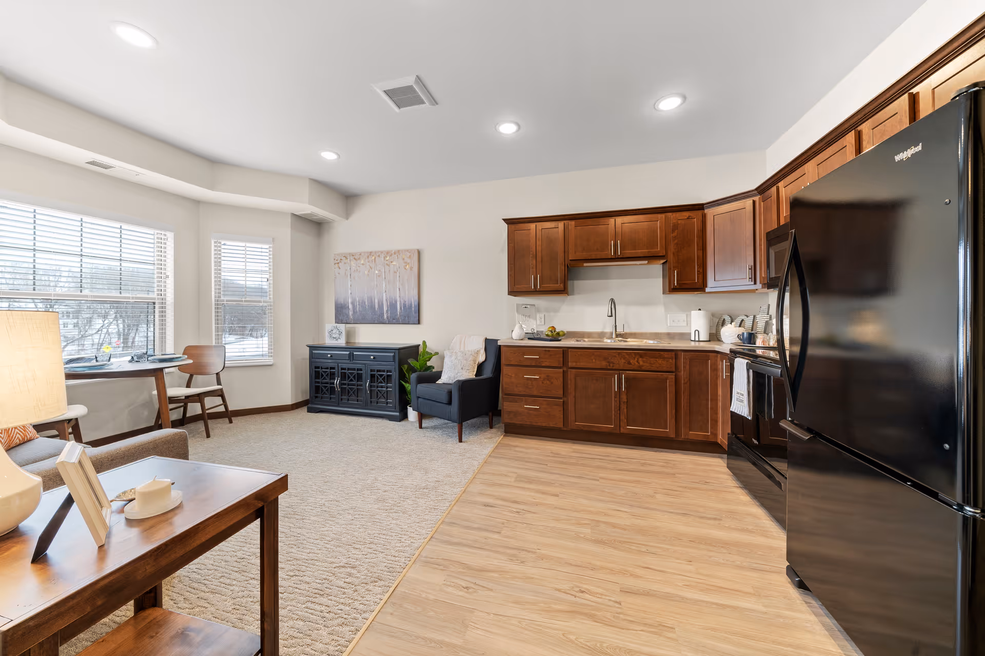 Open-plan senior living apartment interior with a kitchenette, dark wood cabinets and black refrigerator, and a sitting area by large windows.