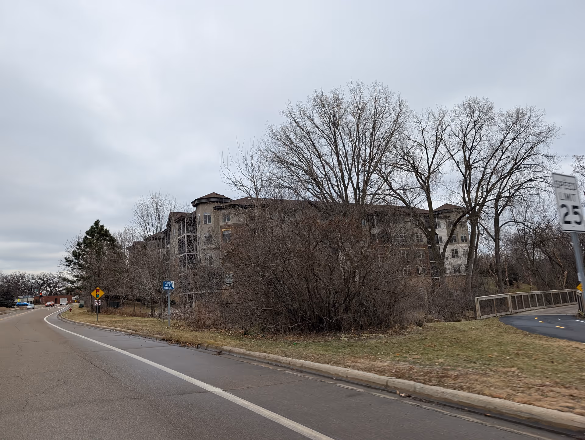 Exterior view of a multi-story senior living facility building partially obscured by leafless trees and bushes, with a road curving alongside it under a cloudy sky.