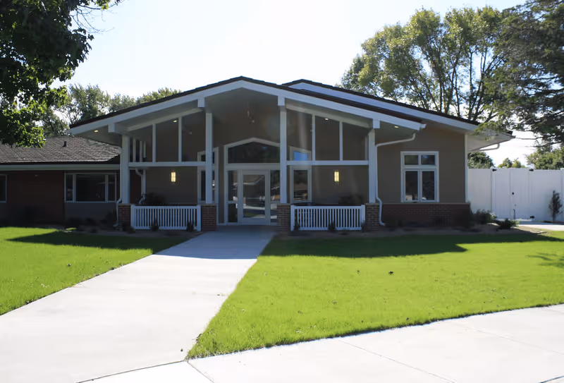 Front exterior view of a single-story senior living facility building with a peaked roof, large windows, and a concrete walkway leading to the entrance. The building is surrounded by green grass and trees under a clear sky.