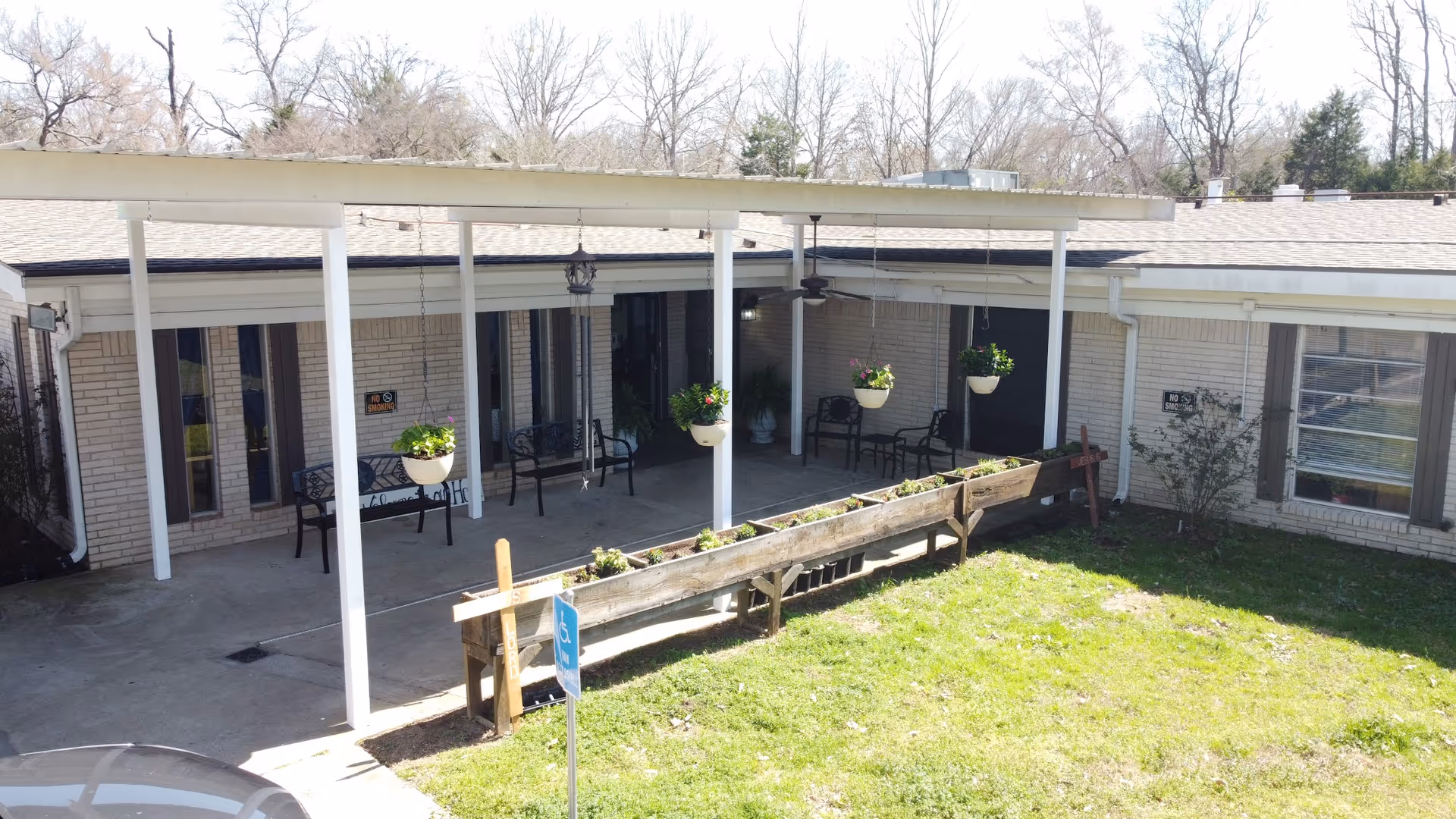 Outdoor patio area at Lakeview Rehabilitation and Healthcare Center with a covered walkway supported by white pillars. There are hanging flower pots with green plants, benches along the wall, and a raised wooden planter box with small plants. The building has light-colored brick walls and several windows. A handicap parking sign is visible near the planter box.