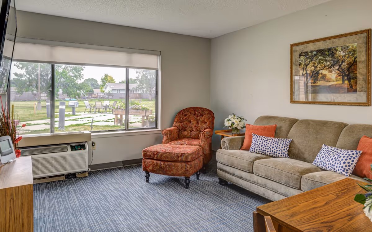 A cozy living room with a large window showing an outdoor garden area. The room features a beige sofa with patterned and solid orange pillows, a red patterned armchair with an ottoman, a wooden side table with a flower arrangement, a wooden cabinet with a TV mounted above it, and a framed picture of a tree-lined path on the wall.