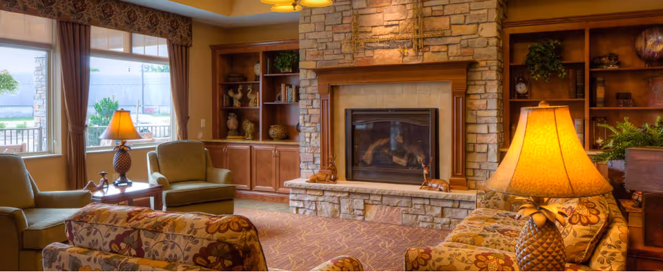 A cozy living room with floral patterned sofas, green armchairs, a stone fireplace with a wooden mantle, built-in wooden shelves with decorative items, and warm lighting from table lamps. Large windows with brown curtains allow natural light to enter the room.