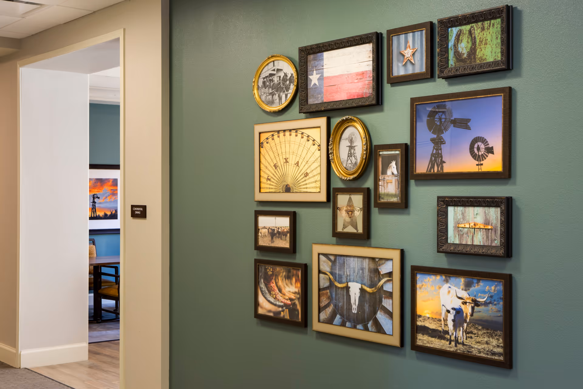 A green wall decorated with multiple framed pictures featuring Texas-themed images such as the Texas flag, windmills, a ferris wheel, a star, a horse, cowboy boots, a cow skull, and cattle. To the left, there is an open doorway leading to a room with a sign that reads 'Continental Dining' and a glimpse of a table and chairs inside.