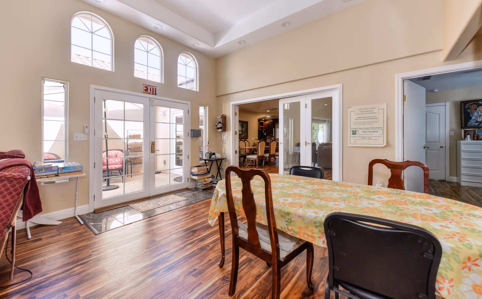 Bright communal dining area with a long table covered in a floral tablecloth, wooden chairs, and glass double doors with an EXIT sign leading outside.