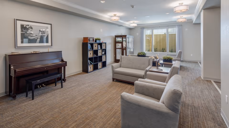 Spacious communal lounge with sofas and armchairs, a piano, shelving, and a large window letting in natural light.