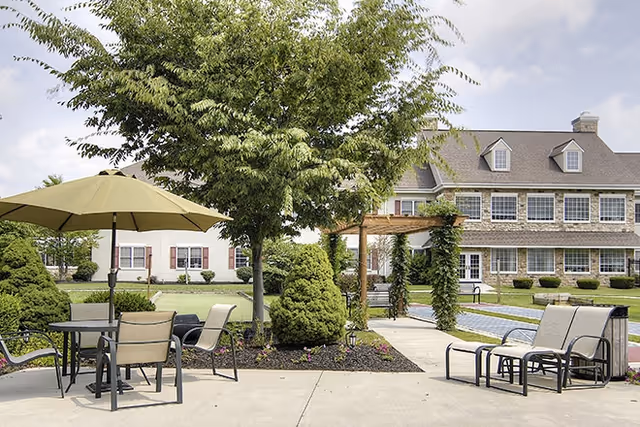 Outdoor patio area at a senior living facility with tables, chairs, and umbrellas surrounded by landscaped greenery and a large tree. In the background, there is a two-story building with multiple windows and a pergola covered in vines.