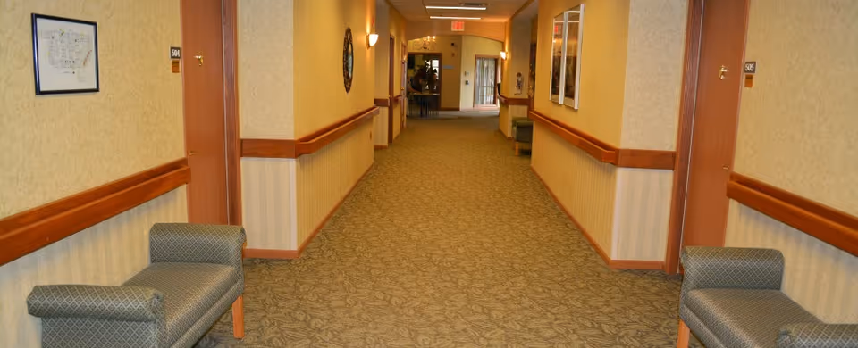 A long, carpeted hallway in a senior living facility with beige walls and wooden trim. There are two upholstered benches on either side near the foreground, doors with room numbers 501 and 506, framed artwork on the walls, and a seating area visible at the far end of the hallway.